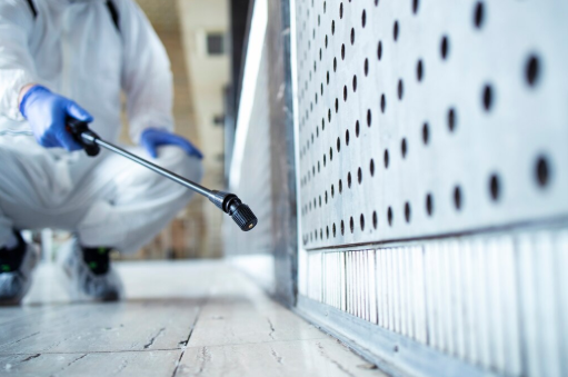 Person in protective suit sprays pesticide on a perforated metal panel indoors; focus on the sprayer and panel.