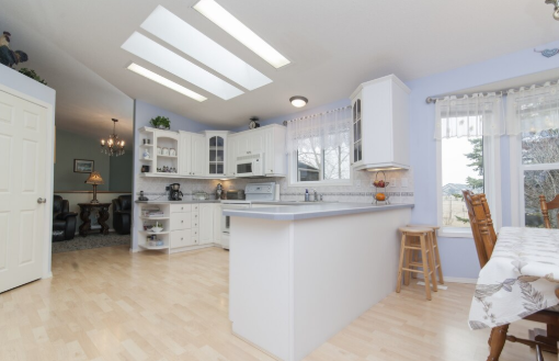 Bright kitchen with white cabinets, island, and skylights. Wooden floor, light blue walls, dining area with chairs on the right.