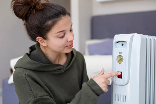 Woman in green hoodie presses a button on a white space heater. Background features a bed with pillows. Calm indoor setting.