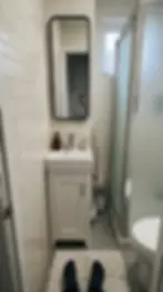 Small bathroom with white tiles, a sink and mirror. A reflection shows someone holding a phone. Rug and black socks visible on floor.