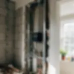 Exposed wall with plumbing and wiring, digital meter at center. Sunlit window with potted plant and bottles in the background.