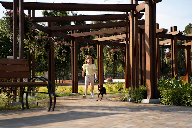 Girl in yellow shirt running with small dog under wooden pergola, surrounded by hanging plants, on a sunny day in a park.