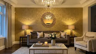 Elegant living room with a velvet sofa, ornate mirror, and chandeliers. Warm lighting enhances gold walls. Candlelit coffee table with flowers.