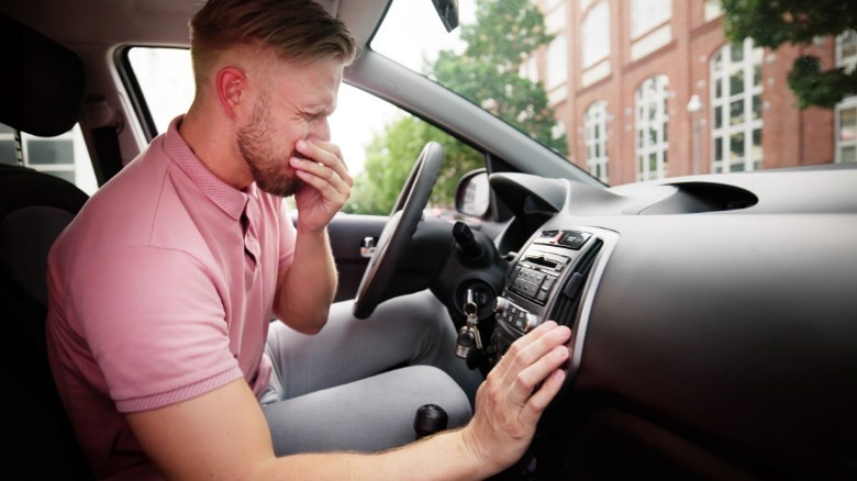 Man in a car, covering his nose, reacts to an unpleasant smell. He's wearing a pink shirt. The car's interior and a building outside are visible.