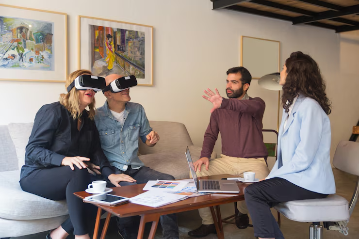 Four people in an office; two wear VR headsets, one gestures animatedly. Documents and a laptop on the table, colorful art on walls.