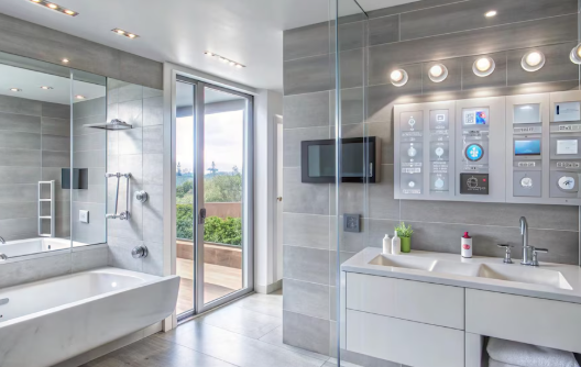 Modern bathroom with gray tiles, large mirror, bathtub, and a digital control panel near the sink. Glass door opens to a green view.
