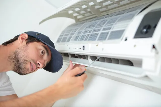 Technician wearing a cap uses a screwdriver to repair a wall-mounted air conditioner. Focused expression, white background.