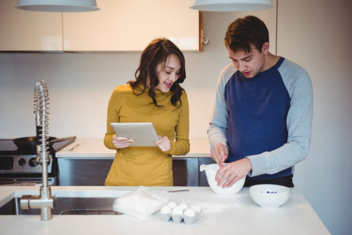 A woman in yellow holds a tablet, smiling as a man in blue mixes ingredients in a white bowl. They're in a modern kitchen with eggs nearby.