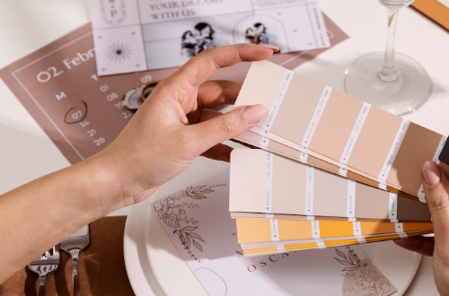 Hands holding paint swatches above a plate, with a calendar and dine setting in the background. Notable colors are peach and muted tones.