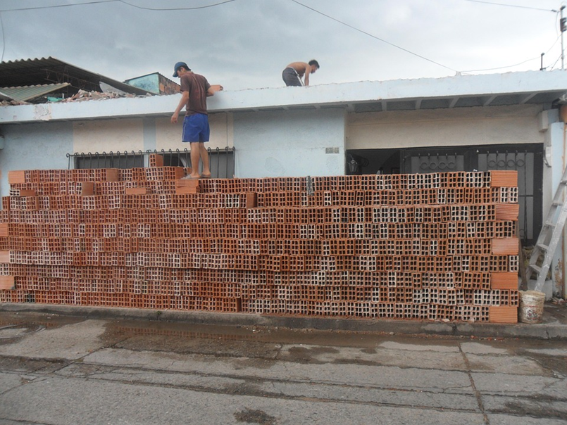 Men stack bricks in front of a blue house. Overcast sky above. One man climbs a ladder. Construction work in progress.