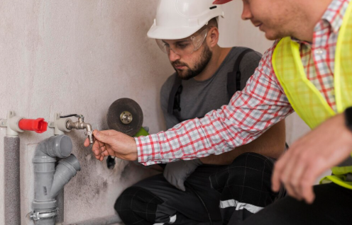Two workers in protective gear adjust a pipe valve; one wears a white helmet, the other a yellow vest. Neutral expression, indoor setting.