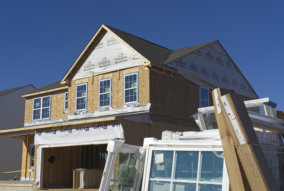 House under construction with visible wooden frames and windows. Clear blue sky in the background, construction materials in front.