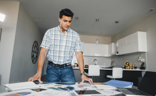 Man in plaid shirt reviewing documents on a table in a modern kitchen, looking focused. Bright lighting, white cabinets in the background.