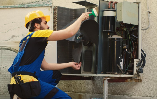 A worker in blue overalls and a yellow hard hat inspects an air conditioning unit outside, using a brush. The setting is sunny and industrial.