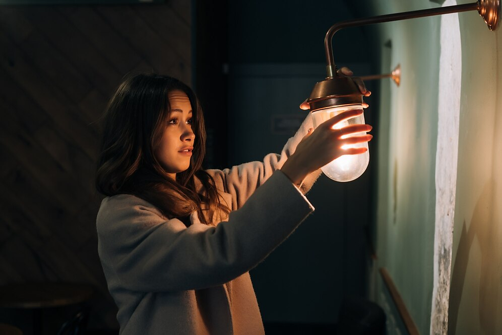 Woman adjusts wall lamp, illuminating dim room with wood-paneled walls. She wears a light coat, expression focused, light casts warm glow.