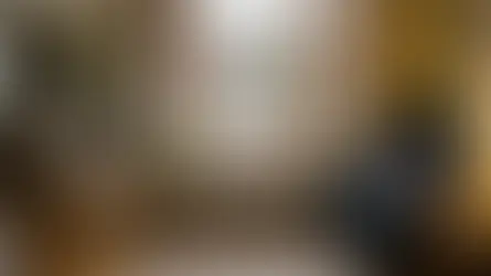 A detailed photograph captures a cozy, traditional British living room centered on a large, multi-paned wooden-framed window with a white column radiator below. On the windowsill rest a potted fern, a coffee mug, and a stack of books, looking out onto a rain-slicked street and brick houses. To the left, a comfortable sofa and built-in bookshelves are visible, while to the right, a blue wingback armchair with a tartan blanket sits next to a side table with a lit lamp. Wide-plank wood flooring and a rug complete the warm interior.