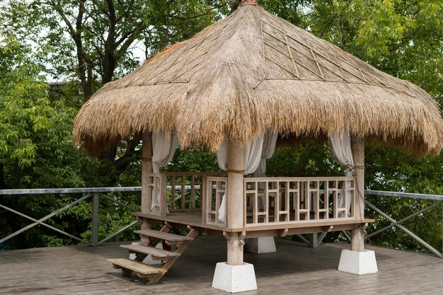 Thatched-roof gazebo with white curtains on a wooden deck surrounded by lush green trees, evoking a serene, tropical ambiance.