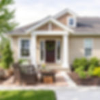 Cozy porch with wicker chairs and table, floral wreath on brown door, green shrubs, tidy garden, and sunny, calm ambiance.