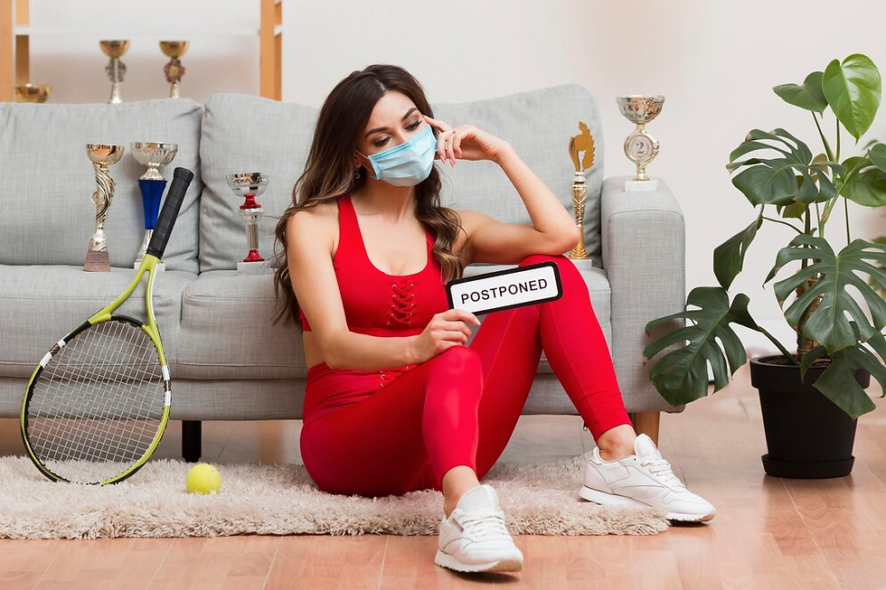 Woman in red sportswear and mask sits on floor holding "Postponed" sign. Tennis racket and trophies nearby. Pensive mood, indoor setting.