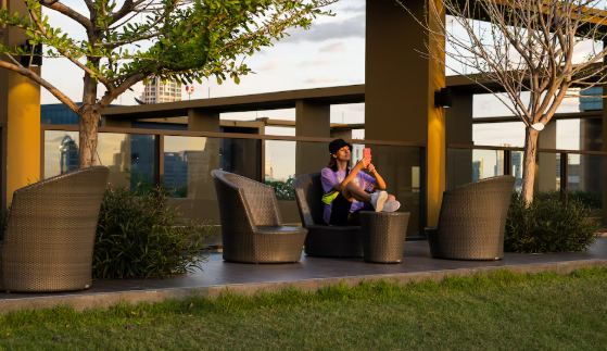 Person sitting on outdoor patio chair using a smartphone, surrounded by trees and wicker furniture, with cityscape in the background.