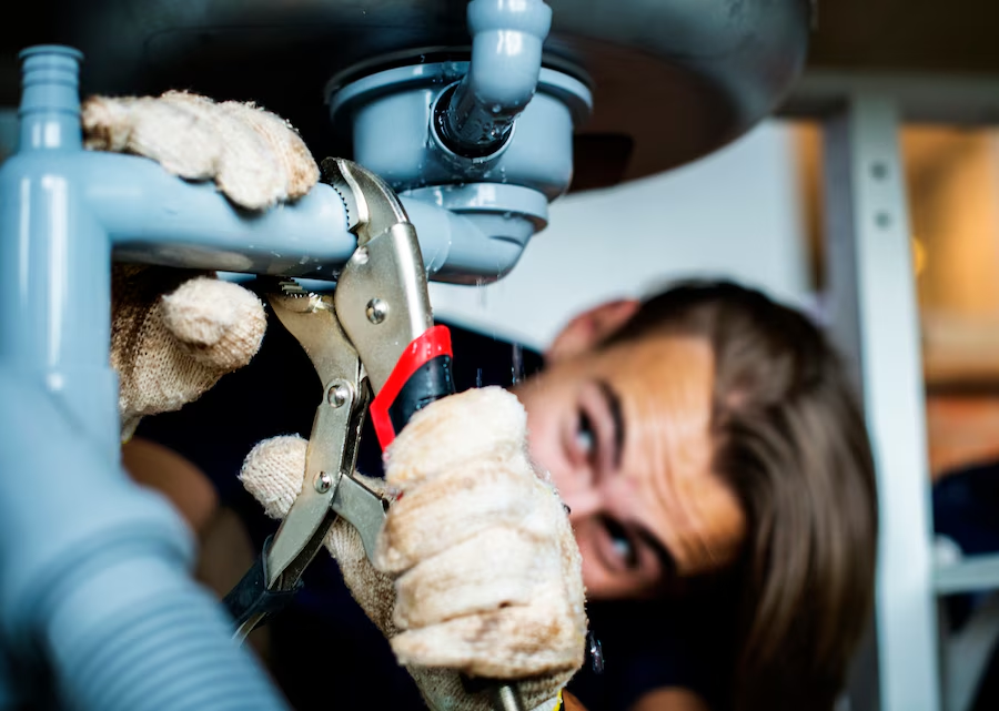 A person uses a wrench to fix a gray pipe under a sink. They wear gloves, focused expression, with blurred background.