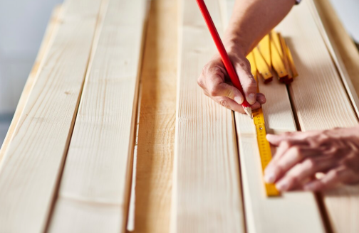 Hands measuring wood planks with a yellow ruler and red pencil in a workshop setting. The wood is light-colored and neatly arranged.