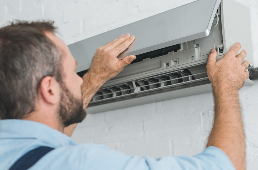 Man adjusts an air conditioner cover on a white brick wall. He wears a blue shirt, focusing calmly on the task. No visible text.