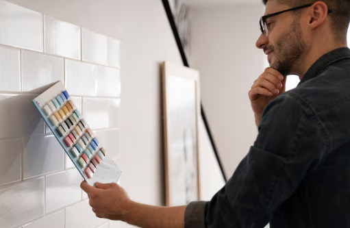 Man in glasses examines paint swatches on a tiled wall, thoughtful expression. Bright, neutral interior with framed artwork in background.