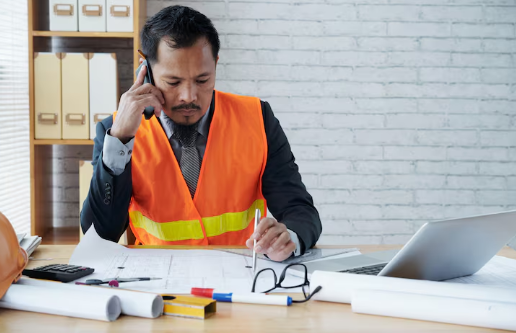 Man in orange safety vest on phone, reviewing blueprints at desk with laptop, glasses, and calculator. White brick wall background.