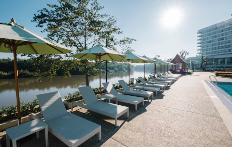 Lounge chairs with green umbrellas by a riverside pool, sunlight glinting, modern building in the background. Serene and sunny atmosphere.