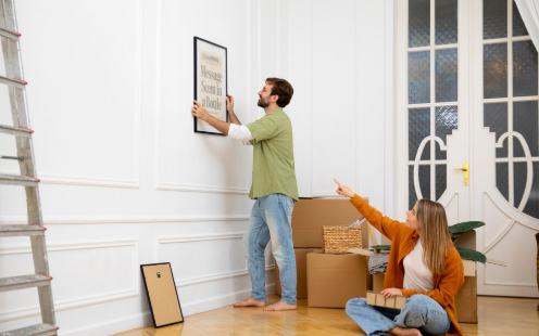 Man hangs framed art on white wall, woman sitting on floor with box points at it. Ladder, cardboard boxes, and door in background. Cozy mood.