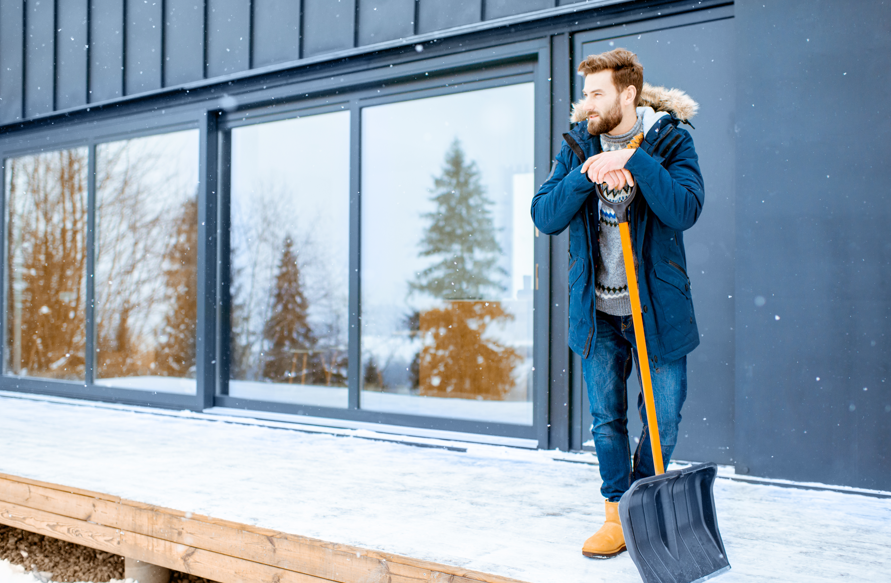 Man in blue coat holds a snow shovel on a snowy deck, in front of a modern building with large windows, with trees in the background.
