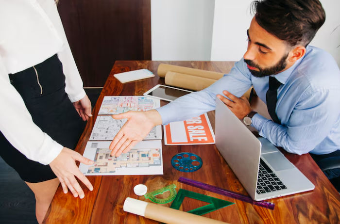 Man and woman discussing blueprints at a wooden table. Man extends hand for a handshake. "For Sale" sign and laptop visible.