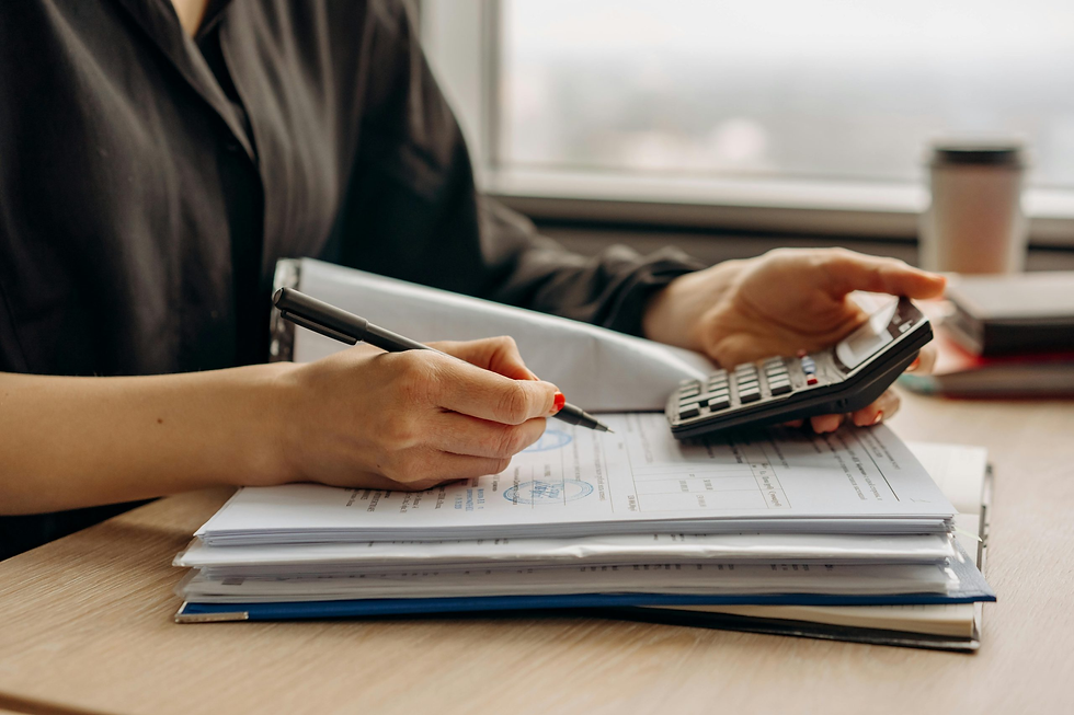 Person in black shirt calculates with a pen and calculator over documents on a wooden desk. A window view and coffee cup are visible.