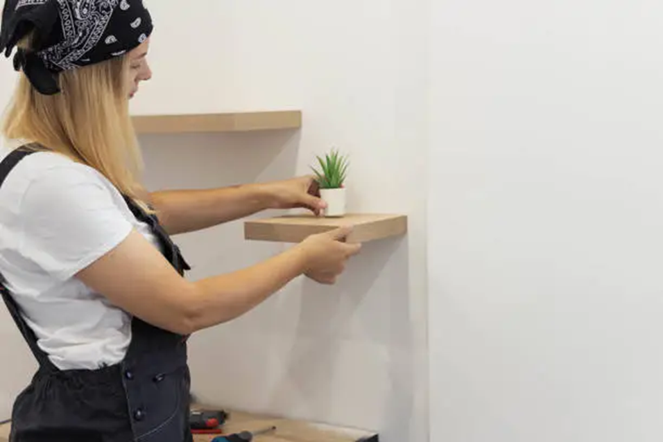 Woman with bandana assembles wooden shelves on a white wall, placing a small plant. Tools are on the table below.