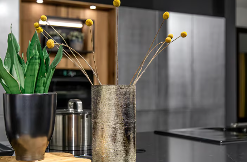Modern kitchen with sleek gray countertops, a metallic vase with yellow flowers, and a black pot with green leaves. Stainless appliances in back.