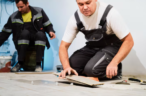Two workers in overalls installing wooden flooring. One kneels, fitting a plank; the other sits in the background. Bright, orderly workspace.