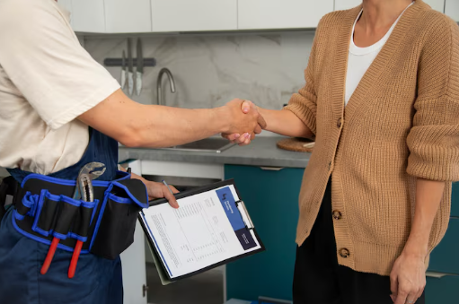 A person in a brown cardigan shakes hands with a worker holding a clipboard in a kitchen. The worker has a tool belt. Professional agreement.