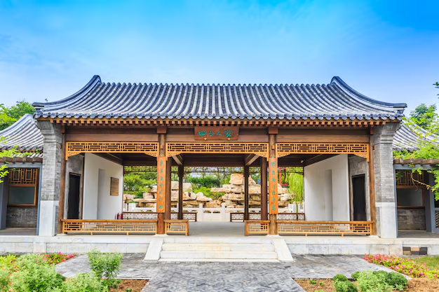 Traditional Asian pavilion with ornate wood details, open to a rock garden. Blue sky, lush greenery, and architectural symmetry enhance the serene setting.