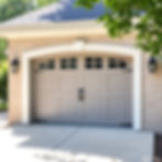 Gray garage door with black handles on a light brick house. Two lantern-style lights flank the door. Green leaves hang from a nearby tree.