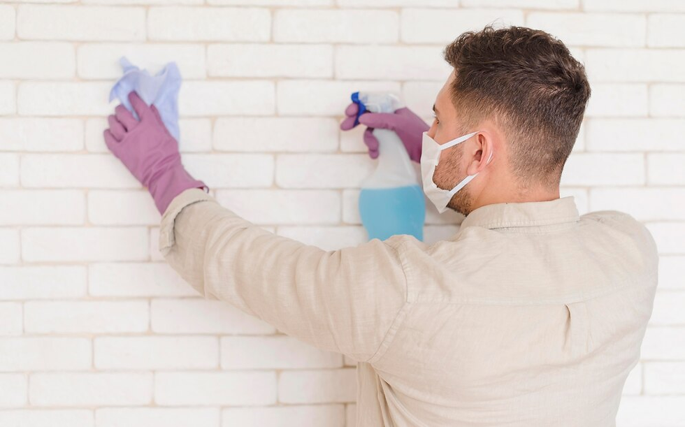 Man in a mask and gloves cleans a white brick wall with blue spray and cloth. Wearing a beige shirt. Focused and hygienic setting.