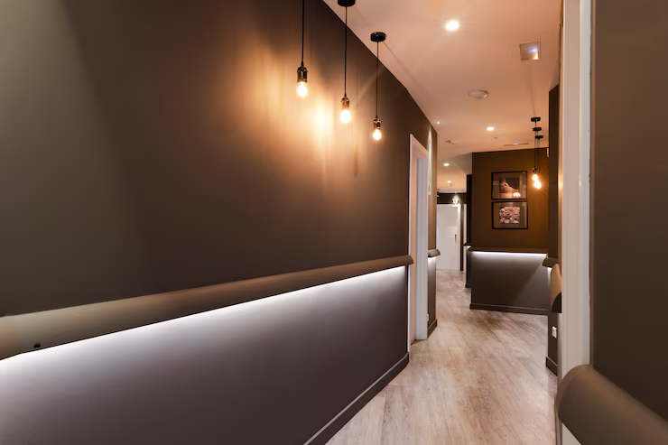 Dimly lit hallway with brown walls, hanging bulbs, and wooden floor. Bright LED strip lighting lines the walls, creating a cozy ambiance.