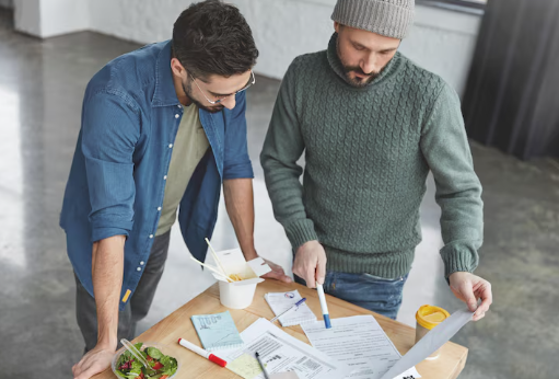 Two men review documents on a table with pens, salad, and takeout. One wears a grey sweater and beanie, the other a blue shirt. Office setting.