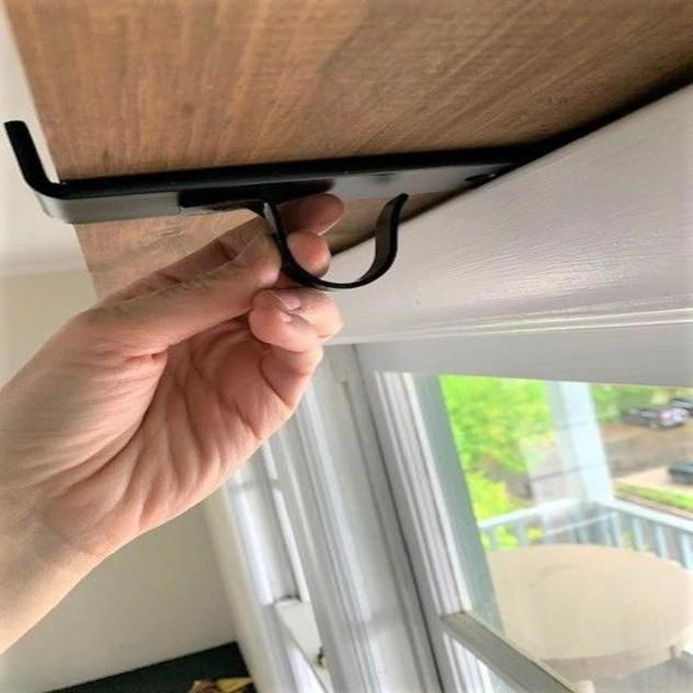 Hand attaching a black metal hook under a wooden shelf above a window, with green foliage visible outside. Bright indoor setting.