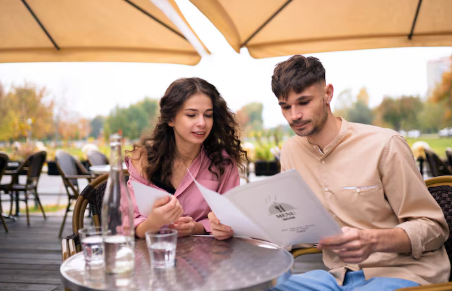 A woman and man sit at an outdoor cafe, reading menus. They appear relaxed, with trees in the background and a warm, calm ambiance.