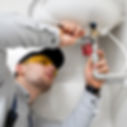 A plumber in gray shirt and cap fixes a pipe with a wrench. Yellow goggles, red valve handles, white wall, and focused expression visible.