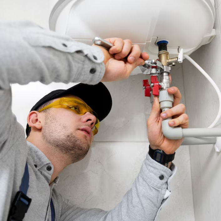 A plumber in gray shirt and cap fixes a pipe with a wrench. Yellow goggles, red valve handles, white wall, and focused expression visible.