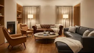 Cozy living room with leather sofas and armchairs. Warm lighting from floor lamps. Curtains, bookshelves, and a modern fireplace create a relaxing vibe.