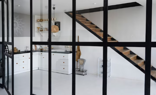 Modern kitchen with white cabinets, exposed black beams, and a wooden staircase. Glass partitions divide the space. Minimalist design.