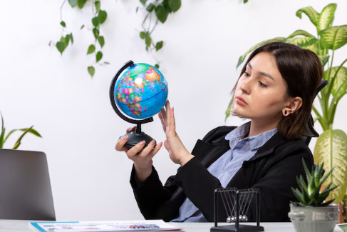 Woman in office attire examines a colorful globe thoughtfully. Plants in the background. Wooden desk with files and small potted plant.